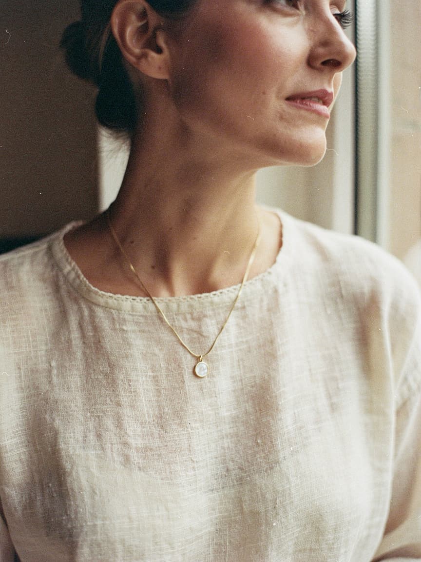 A woman looking out a window, wearing a gold chain with a round mother-of-pearl Memoura pendant